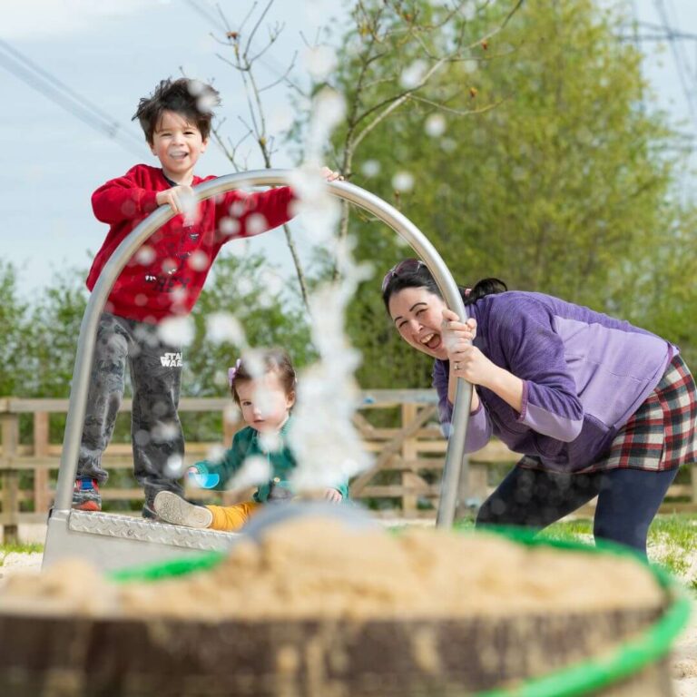 Family laughing at squirty water in a sand play area